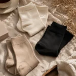 Editorial flat lay composition of bamboo comfort crew socks in ivory, beige, and black arranged on soft linen fabric with warm sunlight.